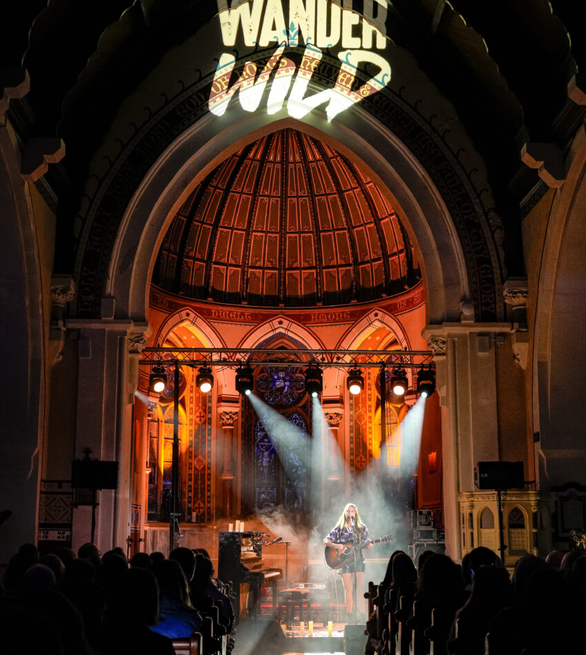 A musician performs on stage in a church-like venue with "Wander Wild" projected above the altar, and an audience seated in pews.