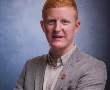A man with short red hair, wearing a grey blazer and light shirt, stands with arms folded against a blue gradient background.