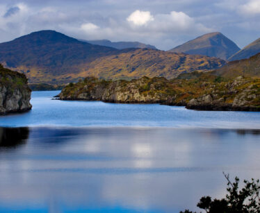 A calm lake surrounded by rocky hills and mountains under a partly cloudy sky, with reflections visible on the water’s surface.