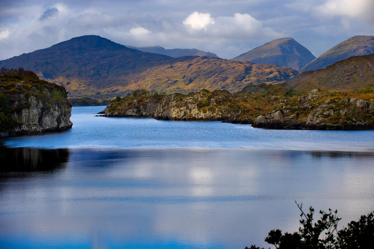 A calm lake surrounded by rocky hills and mountains under a partly cloudy sky, with reflections visible on the water’s surface.