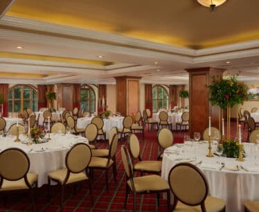 Elegant banqueting hall set for an event with round tables, white tablecloths, floral centrepieces, cushioned chairs, and large windows in the background.