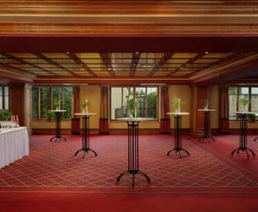 A spacious, carpeted function room with tall cocktail tables arranged in rows, each topped with a small plant. There is a table with drinks set up on the left side. Large windows line the back wall.