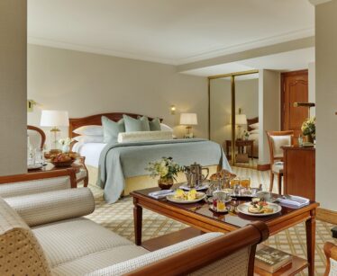 A hotel room with a made bed, armchairs, a wooden table set with breakfast, a mirrored wardrobe, and neutral-coloured decor.