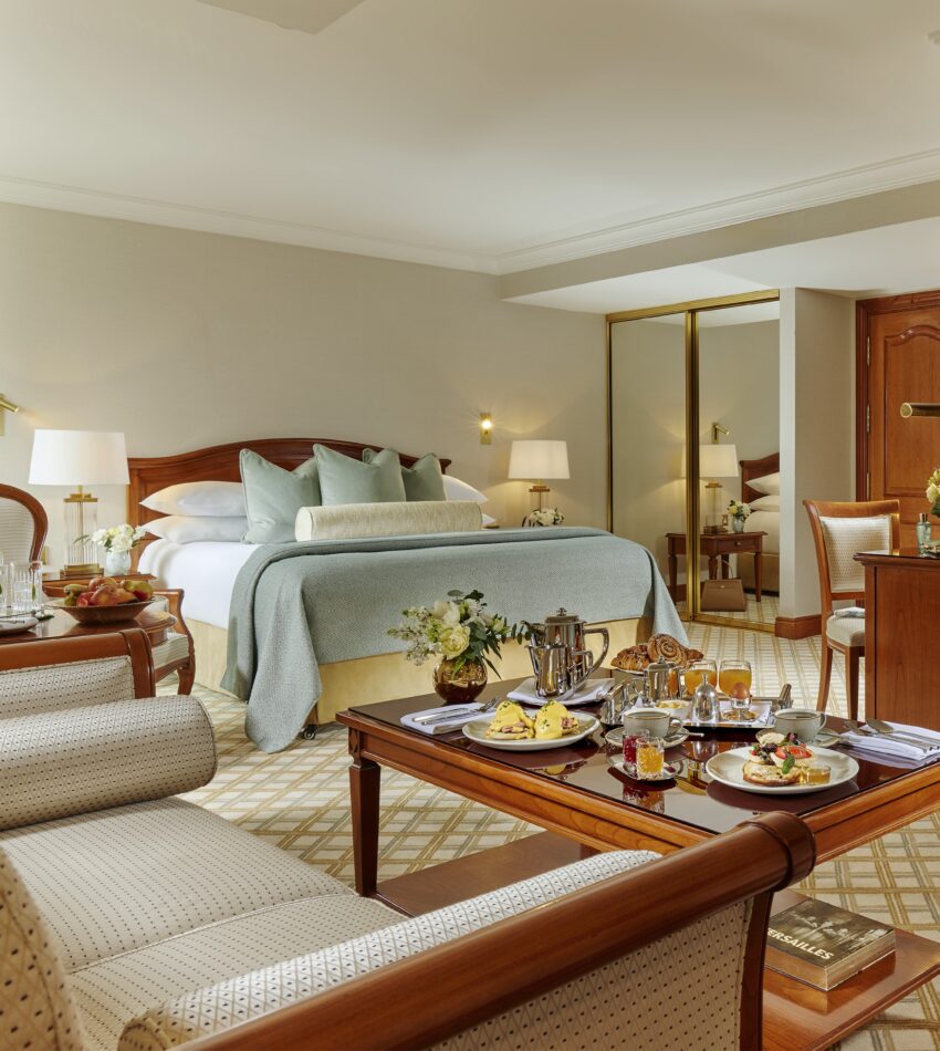 A hotel room with a made bed, armchairs, a wooden table set with breakfast, a mirrored wardrobe, and neutral-coloured decor.