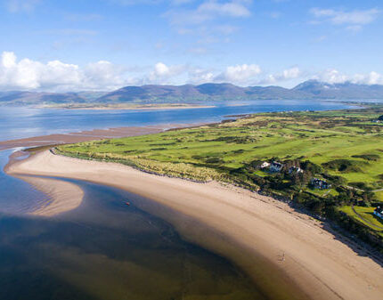 Stunning aerial view of beachfront hotel with lush golf course, serene sandy shores, and mountain vistas.