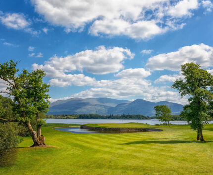 Guests enjoy serene lakeside views, lush green grounds, and mountain vistas under a clear sky at this idyllic hotel.