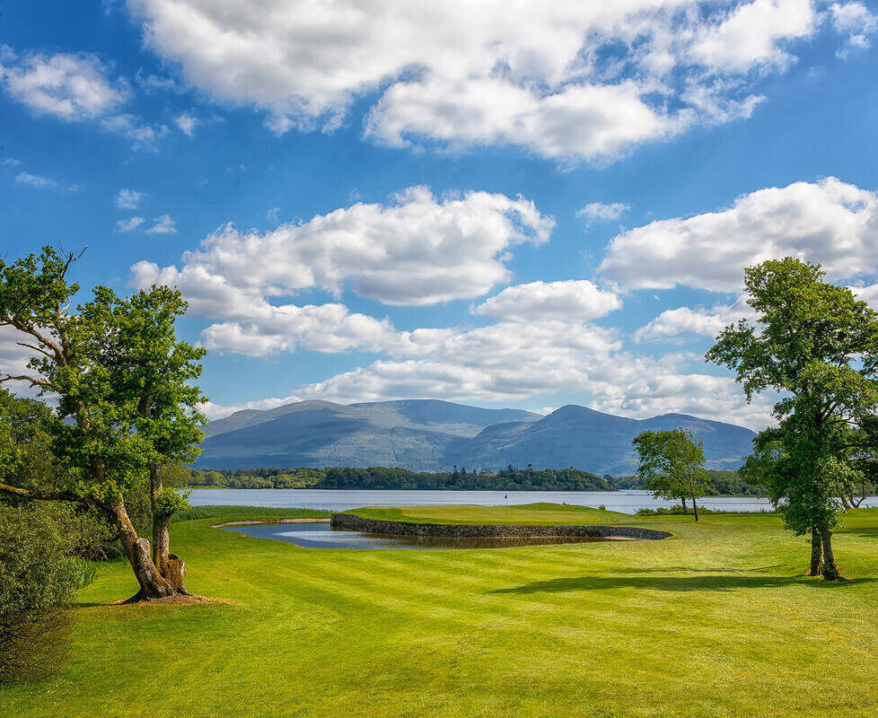 Guests enjoy serene lakeside views, lush green grounds, and mountain vistas under a clear sky at this idyllic hotel.