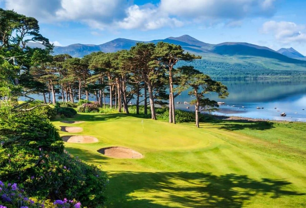 Scenic golf course by lakeside with mountain view, lush greenery, and clear blue sky at luxurious resort.