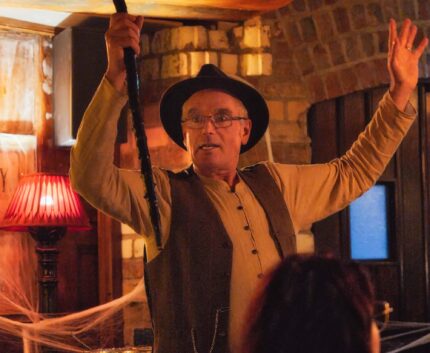 Man in period costume entertains guests in cosy, warmly lit pub, featuring vintage décor and inviting atmosphere.