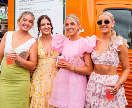 Four smiling women enjoying summer drinks by a vibrant orange food truck at the outdoor café.