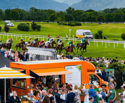 Scenic racecourse view with jockeys on horseback, vibrant crowd, food trucks, and lush mountains in the backdrop.