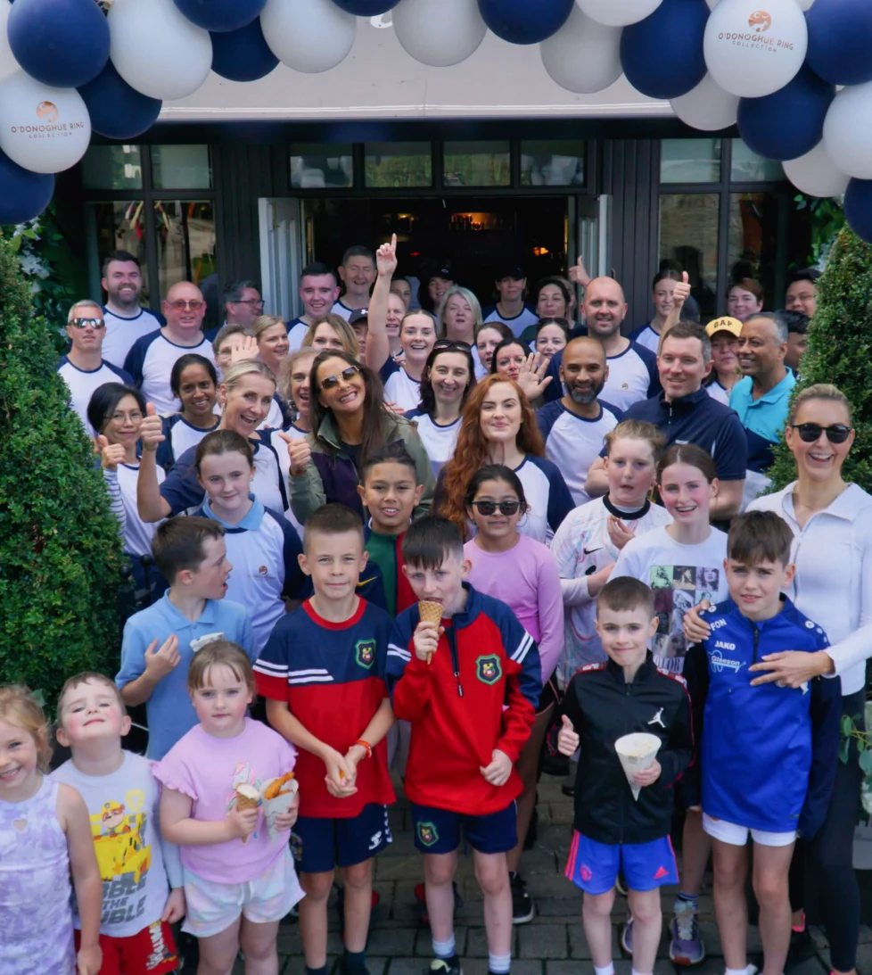 Smiling group of guests and children under navy and white balloon arch at hotel entrance, outdoor greenery, ice creams, welcome event