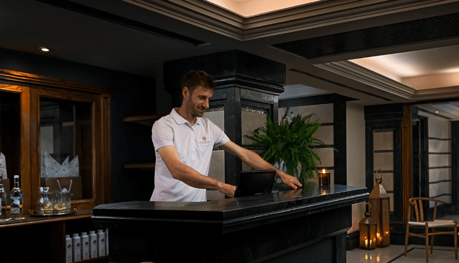 Friendly hotel receptionist at a dark stone check‑in desk using a tablet, warm lobby lighting, decorative plants and candlelit lanterns visible